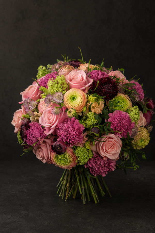 A vibrant, dense floral bouquet featuring pink and purple roses, magenta carnations, light green hydrangeas, and dark burgundy anemones, set against a dark textured studio background.