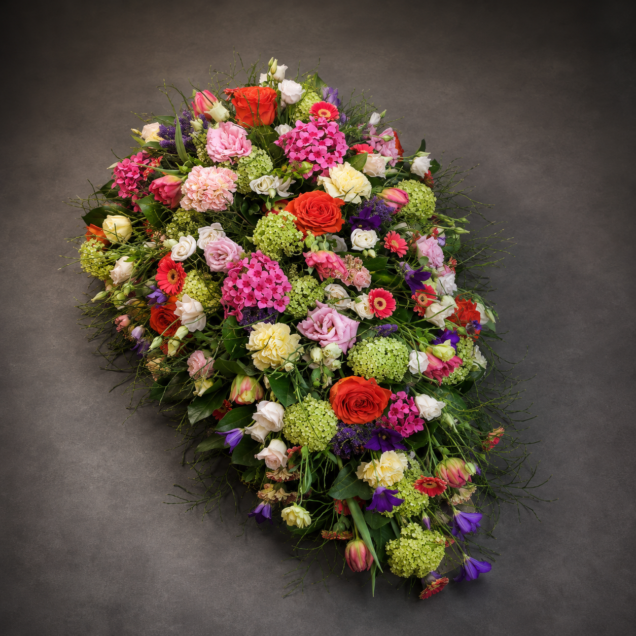 A large, vibrant floral arrangement, likely a casket or catafalque flower bouquet, featuring a dense mix of multicolored flowers including pink, red, yellow, purple, and green, set against a dark gray studio background.