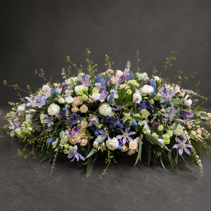 A large, lush floral arrangement featuring white roses, purple lilies, blue clematis, and soft peach roses with green foliage, displayed on a dark gray textured surface against a dark gray background.
