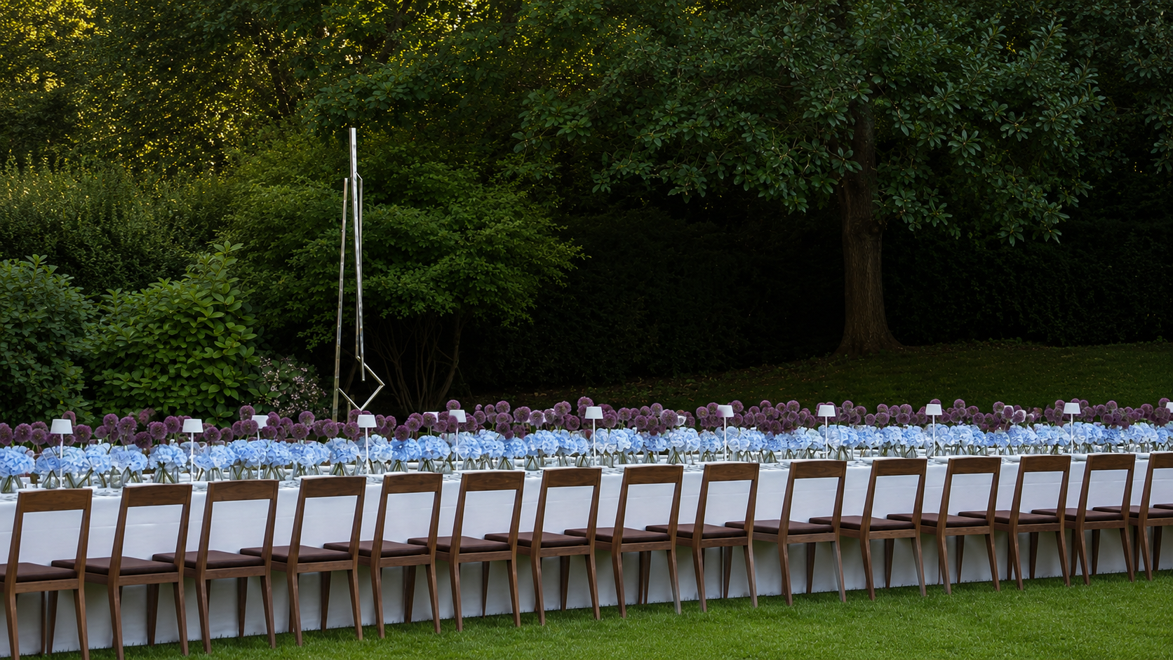 A long outdoor event table set with white tablecloths, brown wooden chairs, and rows of blue and purple flowers, positioned on a green lawn with a backdrop of dark trees.