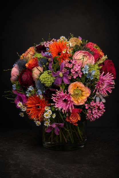 A vibrant, multicolored floral bouquet featuring orange gerbera daisies, pink ranunculus, purple clematis, and blue thistles arranged in a clear glass jar against a dark, textured background.