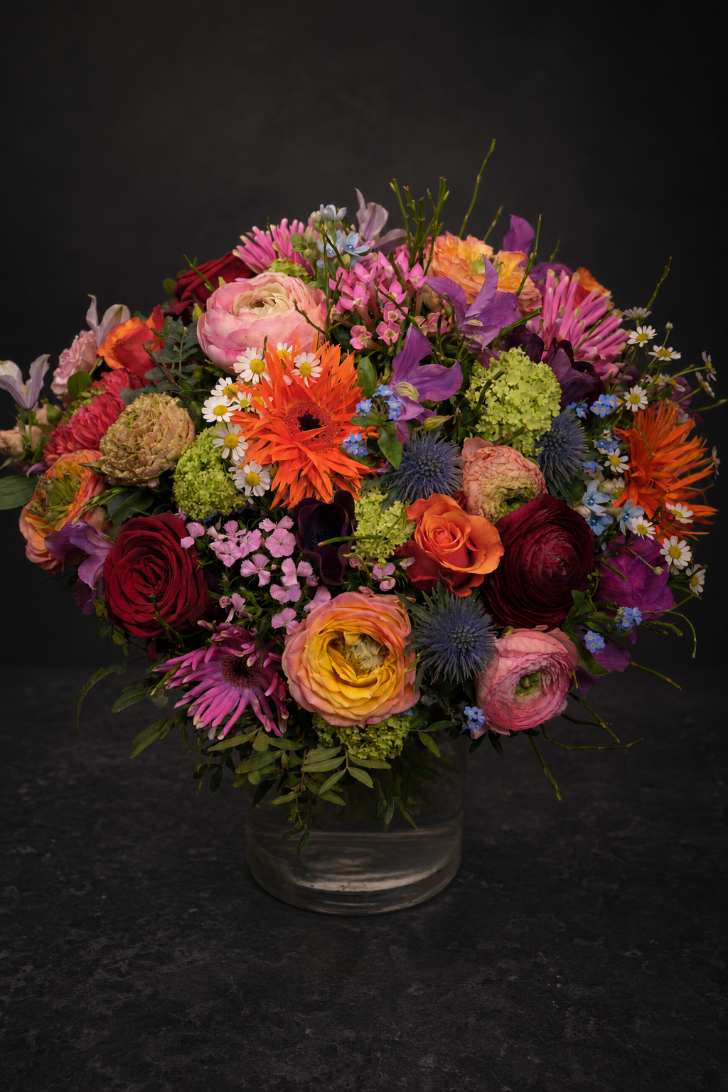 A vibrant, multicolor floral arrangement featuring orange gerbera daisies, pink ranunculus, red and purple roses, and green thistles in a clear glass vase against a dark background.