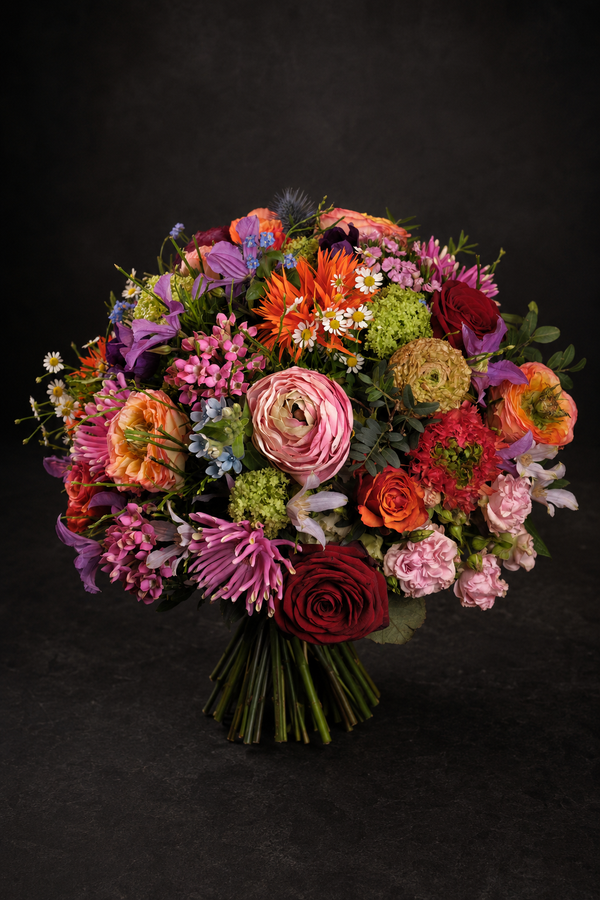 A vibrant, multicolored floral bouquet featuring roses, ranunculus, daisies, and other wildflowers, set against a dark textured studio background.