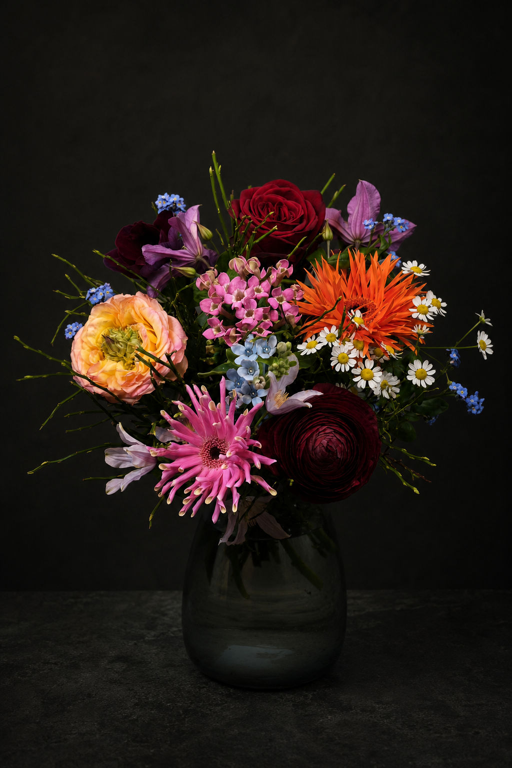 A vibrant mixed flower bouquet in a dark glass vase against a dark, textured background. The bouquet features a variety of colorful flowers including orange daisy-like blooms, pink ranunculus, red roses, purple lilies, and small white daisies.