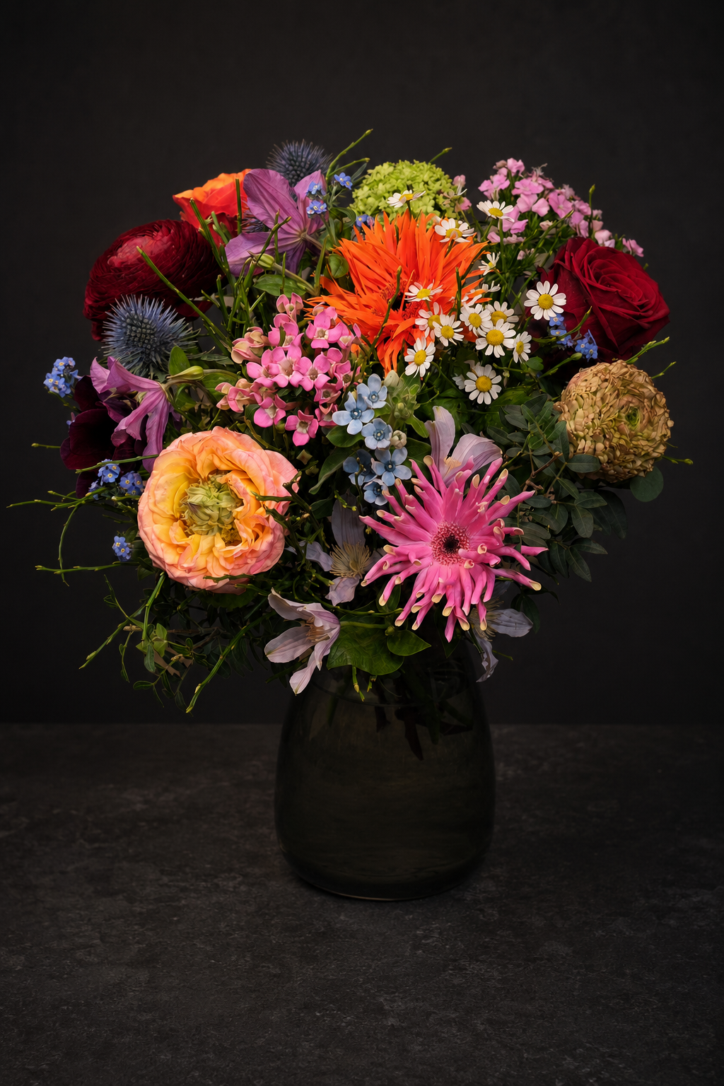 A vibrant, multicolored wildflower bouquet arranged in a dark glass vase against a dark, textured background.