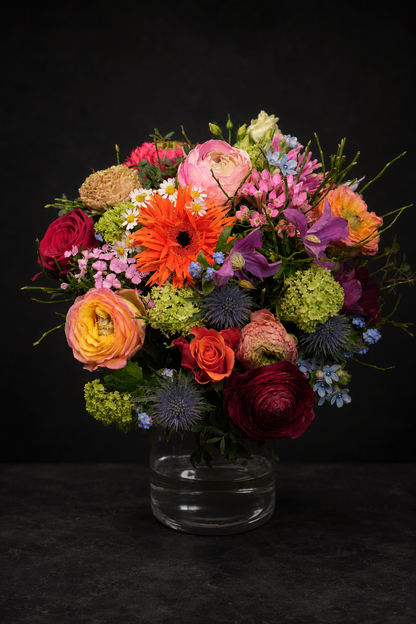 A vibrant, multicolored floral arrangement featuring orange gerbera daisies, pink and yellow ranunculus, purple thistles, and red roses in a clear glass vase against a dark background.
