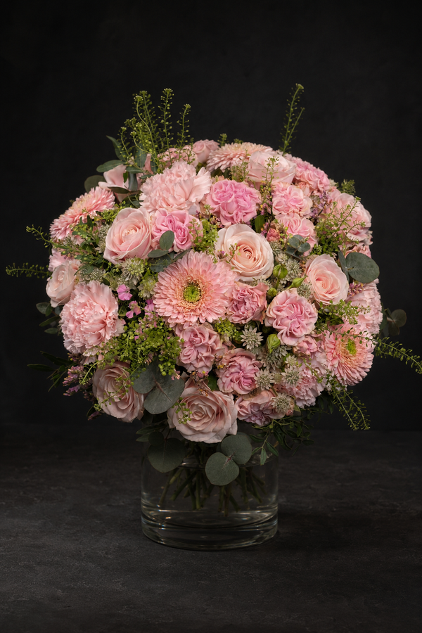 A round floral arrangement of pink roses, chrysanthemums, and green foliage in a clear glass vase, set against a dark black background.