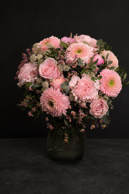 A vibrant floral arrangement of pink roses, pink chrysanthemums, and green foliage in a dark glass vase, set against a solid black background.