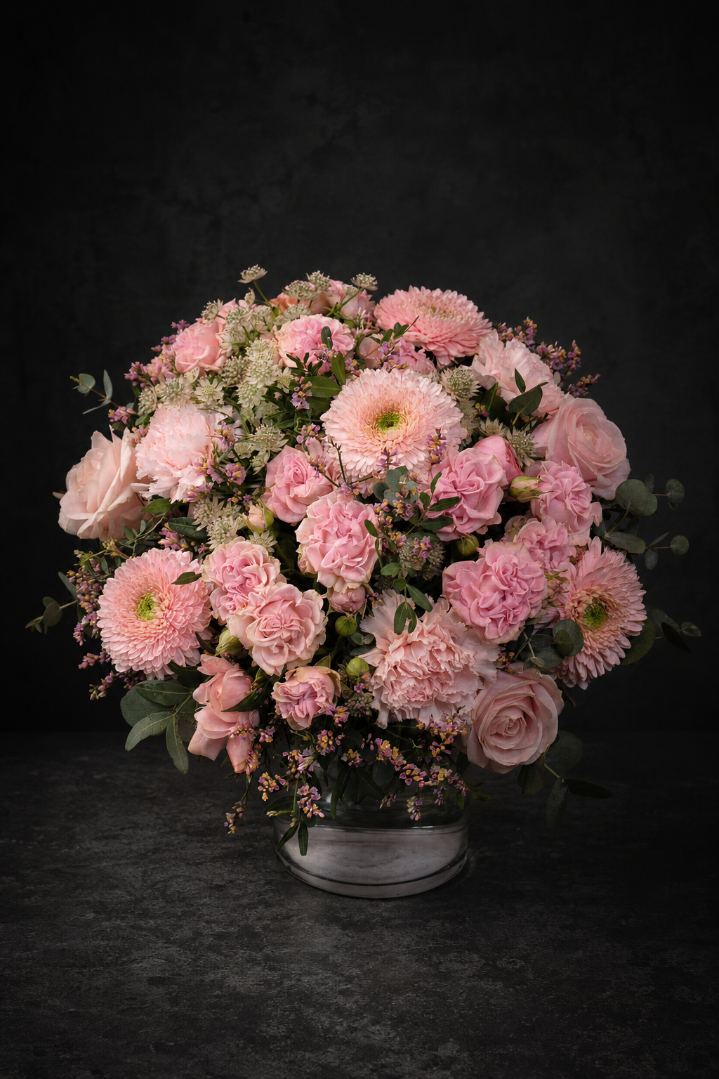 A round floral arrangement of pink roses, chrysanthemums, and carnations with green foliage, presented in a clear glass vase on a dark textured surface against a dark background.