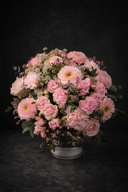 A round floral arrangement of pink roses, chrysanthemums, and carnations with green foliage, presented in a clear glass vase on a dark textured surface against a dark background.