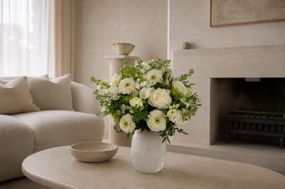 A white ceramic vase filled with a lush arrangement of white flowers and green foliage, set on a round beige stone coffee table in a modern, minimalist living room with a fireplace and sofa.