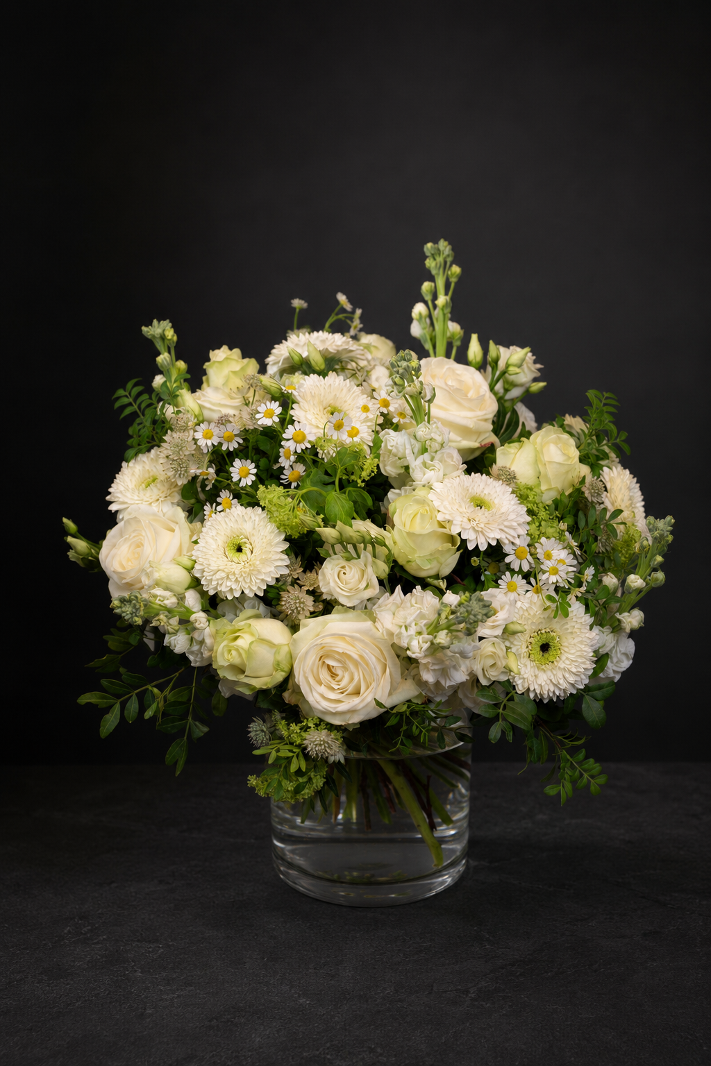 A fresh floral arrangement of white and cream roses, daisies, and green foliage in a clear glass vase, set against a dark black background.