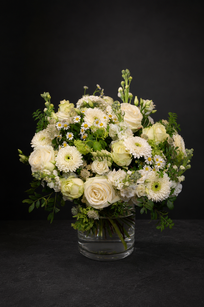 A fresh floral arrangement of white and cream roses, daisies, and green foliage in a clear glass vase, set against a dark black background.