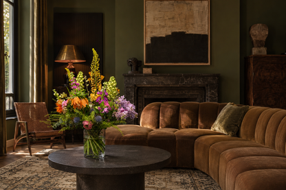 A luxurious curved sectional sofa upholstered in rich brown velvet, styled in a dark, elegant living room with a round stone coffee table, a vibrant floral arrangement, and a dark fireplace.