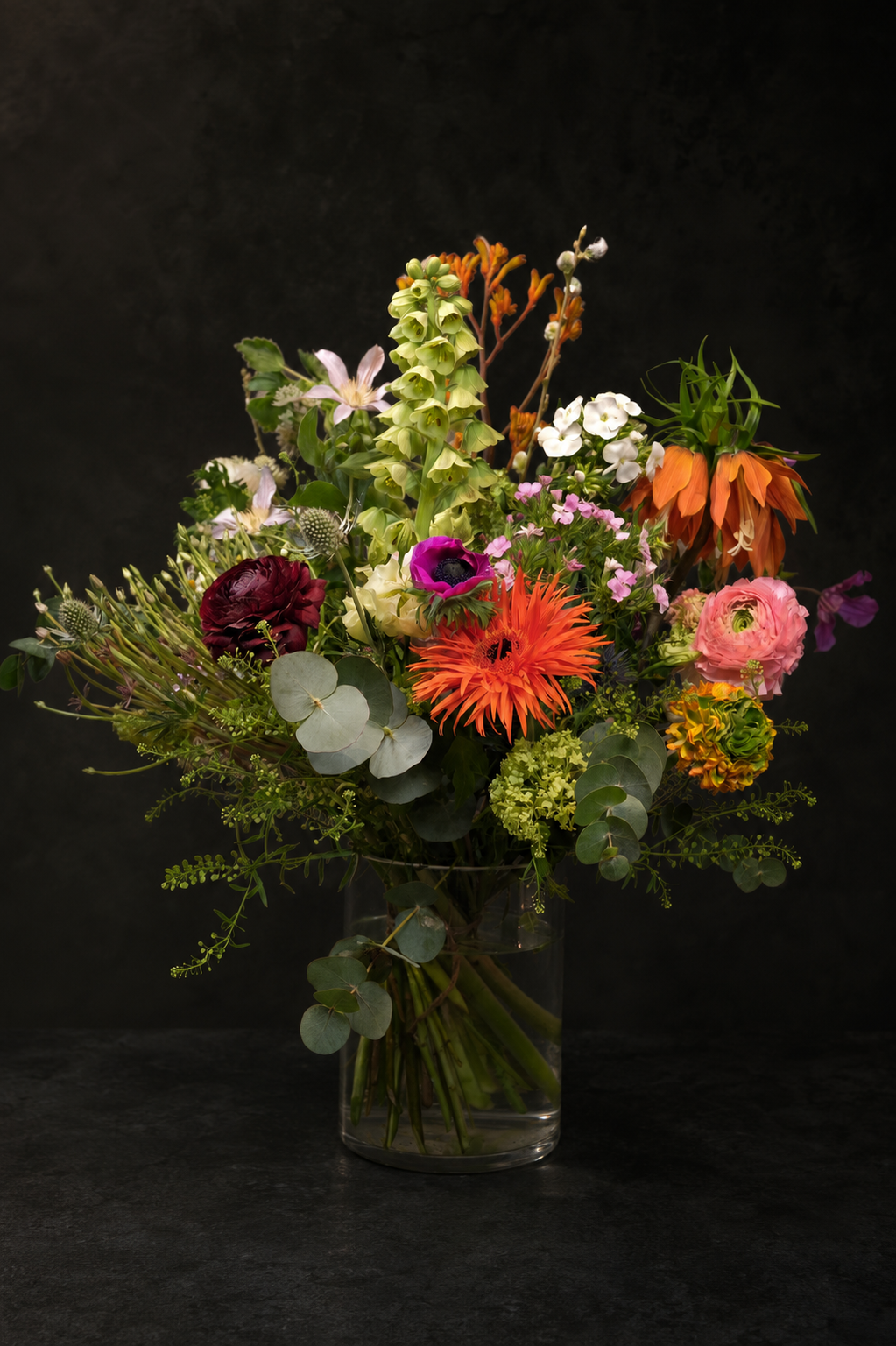 A vibrant mixed floral arrangement in a clear glass vase, featuring orange gerbera daisies, pink ranunculus, purple anemones, and green foliage, set against a dark textured background.