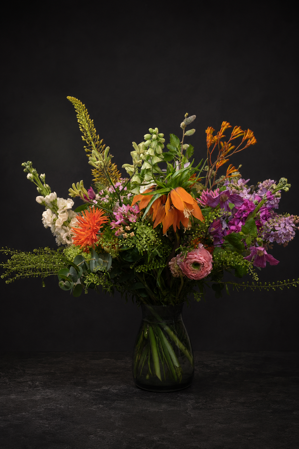 A vibrant mixed flower bouquet in a dark green glass vase, featuring orange, purple, pink, and white flowers with green foliage, set against a dark studio background.