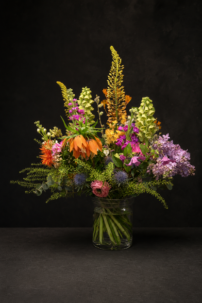 A vibrant, multicolored wildflower bouquet arranged in a clear glass jar, set against a dark, textured studio background.