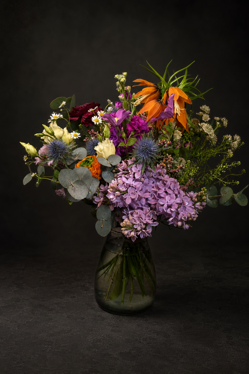 A vibrant mixed flower bouquet in a dark glass vase against a dark studio background. The bouquet features prominent purple hydrangeas, orange lilies, blue thistle spheres, white daisies, and green foliage.