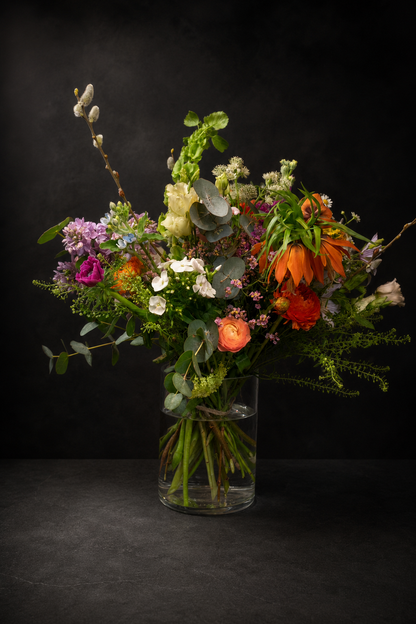 A vibrant mixed floral arrangement in a clear glass vase, featuring orange poppies, pink and purple flowers, green foliage, and eucalyptus, set against a dark textured background.